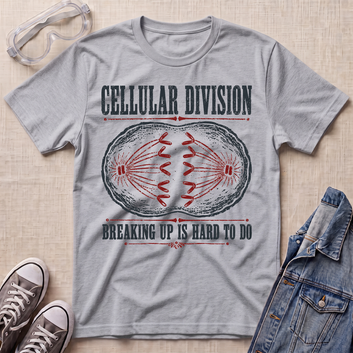A heather gray graphic t-shirt laid flat on a beige textured background, styled with safety glasses, sneakers, and a denim jacket. The t-shirt features a dark red and black graphic of a dividing cell with the text 'CELLULAR DIVISION' and 'BREAKING UP IS HARD TO DO'.
