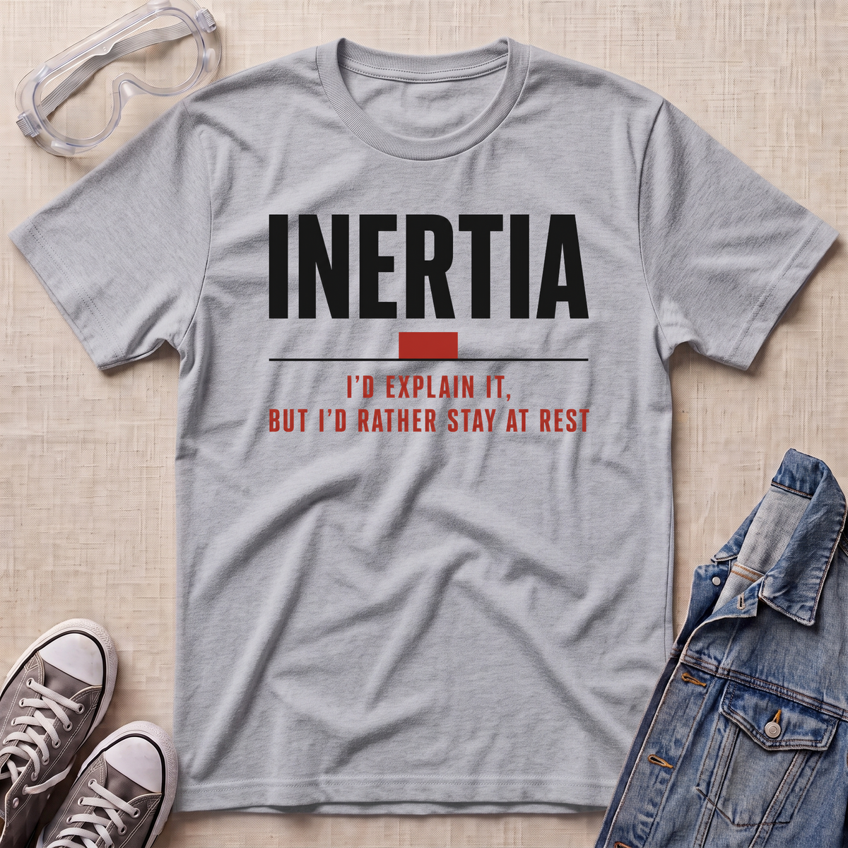 A heather gray graphic t-shirt laid flat on a light wood background, styled with safety goggles, sneakers, and a denim jacket. The t-shirt features the word 'INERTIA' in bold black letters with a red block and red text underneath reading 'I'D EXPLAIN IT, BUT I'D RATHER STAY AT REST'.