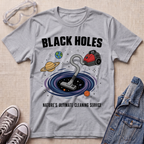 A sport grey graphic t-shirt laid flat on a textured fabric background, styled with safety goggles, sneakers, and a denim jacket. The t-shirt features a cartoon illustration of a vacuum cleaner falling into a black hole surrounded by planets, with the text 'BLACK HOLES' and 'NATURE'S ULTIMATE CLEANING SERVICE'.
