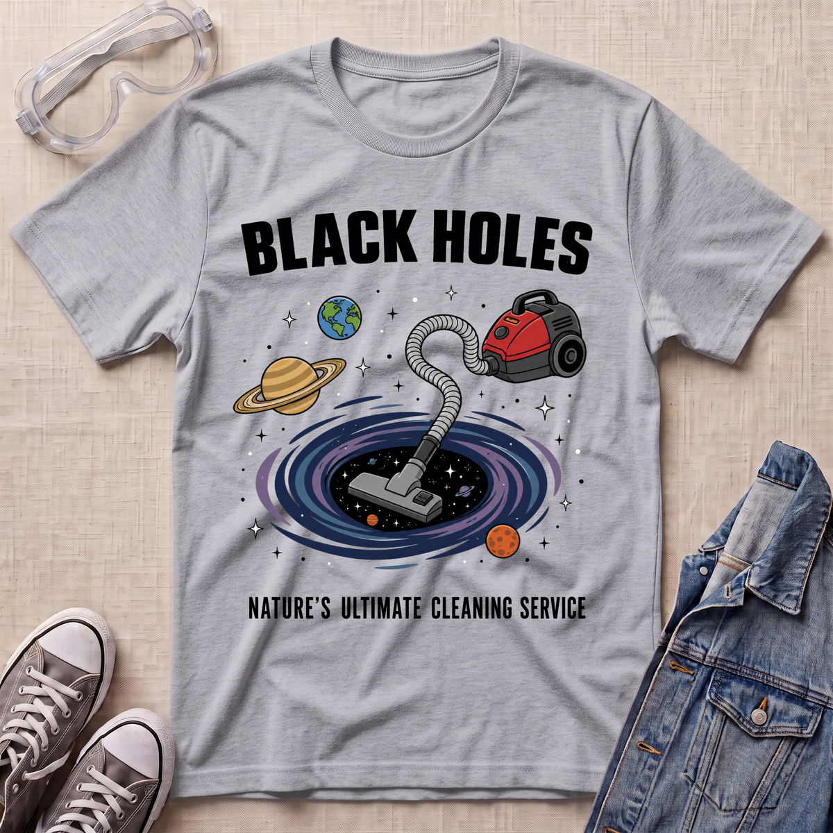 A sport grey graphic t-shirt laid flat on a textured fabric background, styled with safety goggles, sneakers, and a denim jacket. The t-shirt features a cartoon illustration of a vacuum cleaner falling into a black hole surrounded by planets, with the text 'BLACK HOLES' and 'NATURE'S ULTIMATE CLEANING SERVICE'.