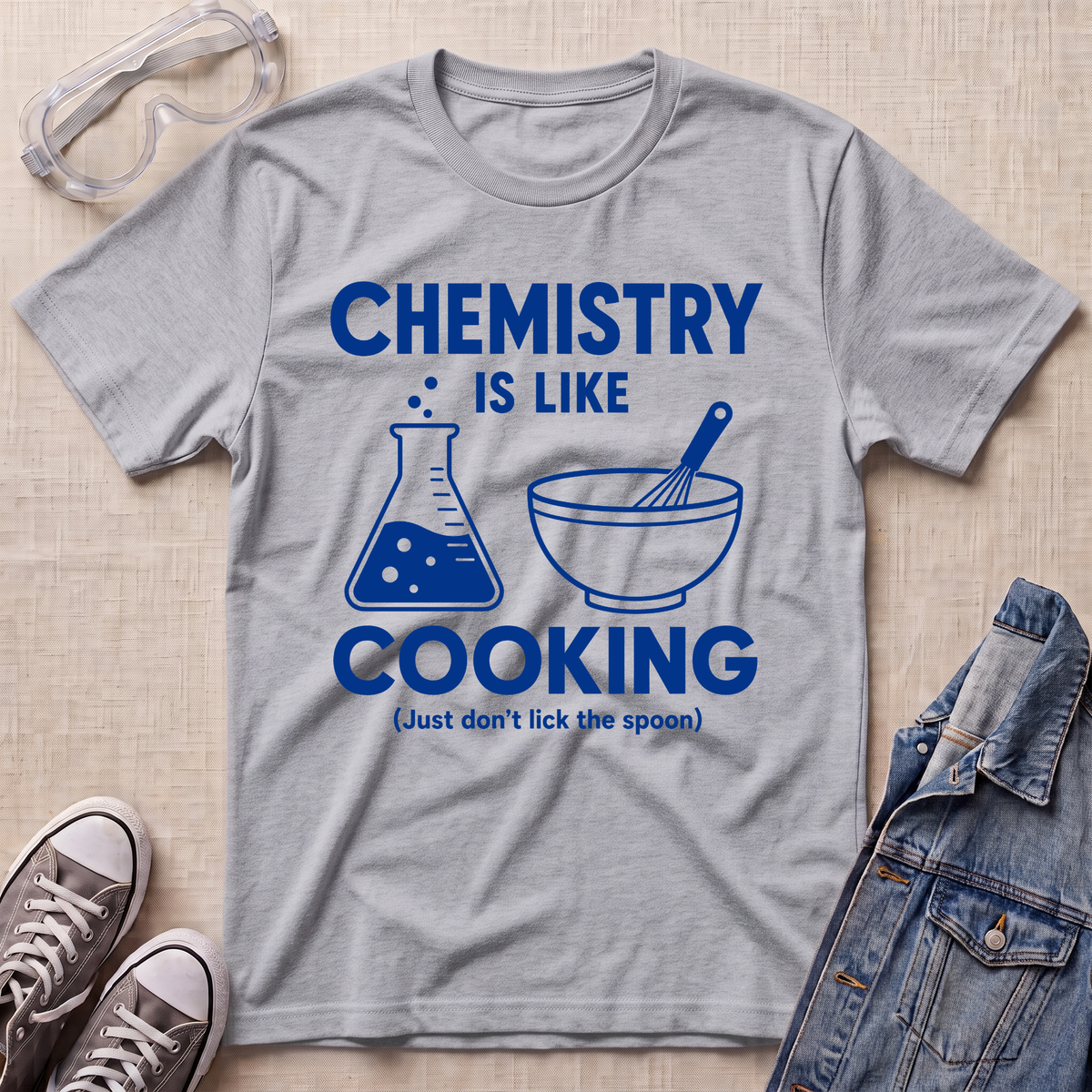 A heather gray short-sleeve t-shirt with a blue graphic print that reads 'CHEMISTRY IS LIKE COOKING (Just don't lick the spoon)' with a beaker and bowl illustration, styled in a flat lay with safety goggles, sneakers, and denim on a light wood background.