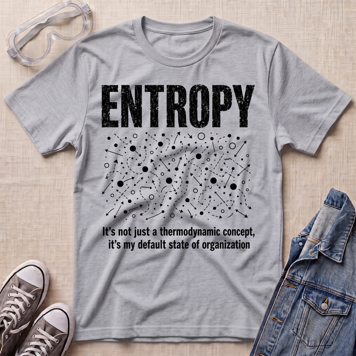 A heather gray graphic t-shirt laid flat on a beige fabric background, styled with safety goggles, gray sneakers, and a denim jacket. The t-shirt features a white distressed graphic of the word 'ENTROPY' above a entropy pattern, with the text 'It's not just a thermodynamic concept, it's my default state of organization' below.