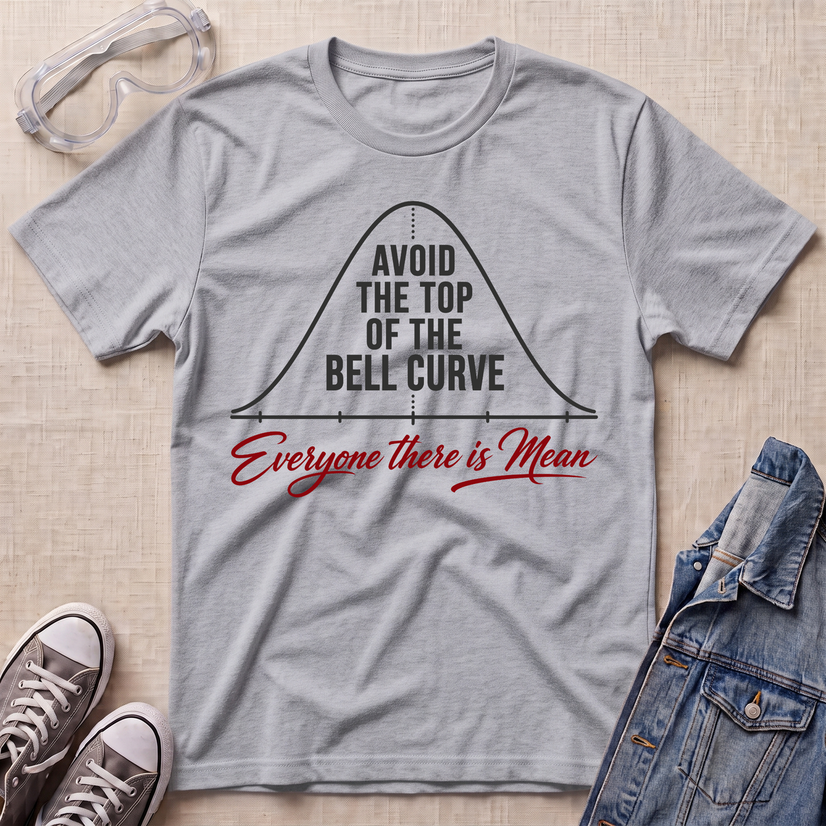 A heather gray graphic t-shirt laid flat on a light wood background, styled with safety goggles, gray sneakers, and a denim jacket. The t-shirt features a black bell curve graphic with the text 'AVOID THE TOP OF THE BELL CURVE' and red script text reading 'Everyone there is Mean'.