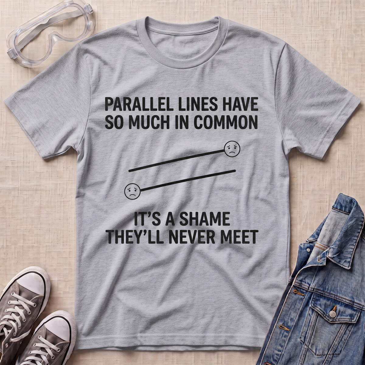 A heather gray graphic t-shirt laid flat on a beige textured surface, surrounded by props including safety goggles, sneakers, and a denim jacket. The t-shirt features a black printed graphic of two parallel sad faces with the text 'PARALLEL LINES HAVE SO MUCH IN COMMON' and 'IT'S A SHAME THEY'LL NEVER MEET'.
