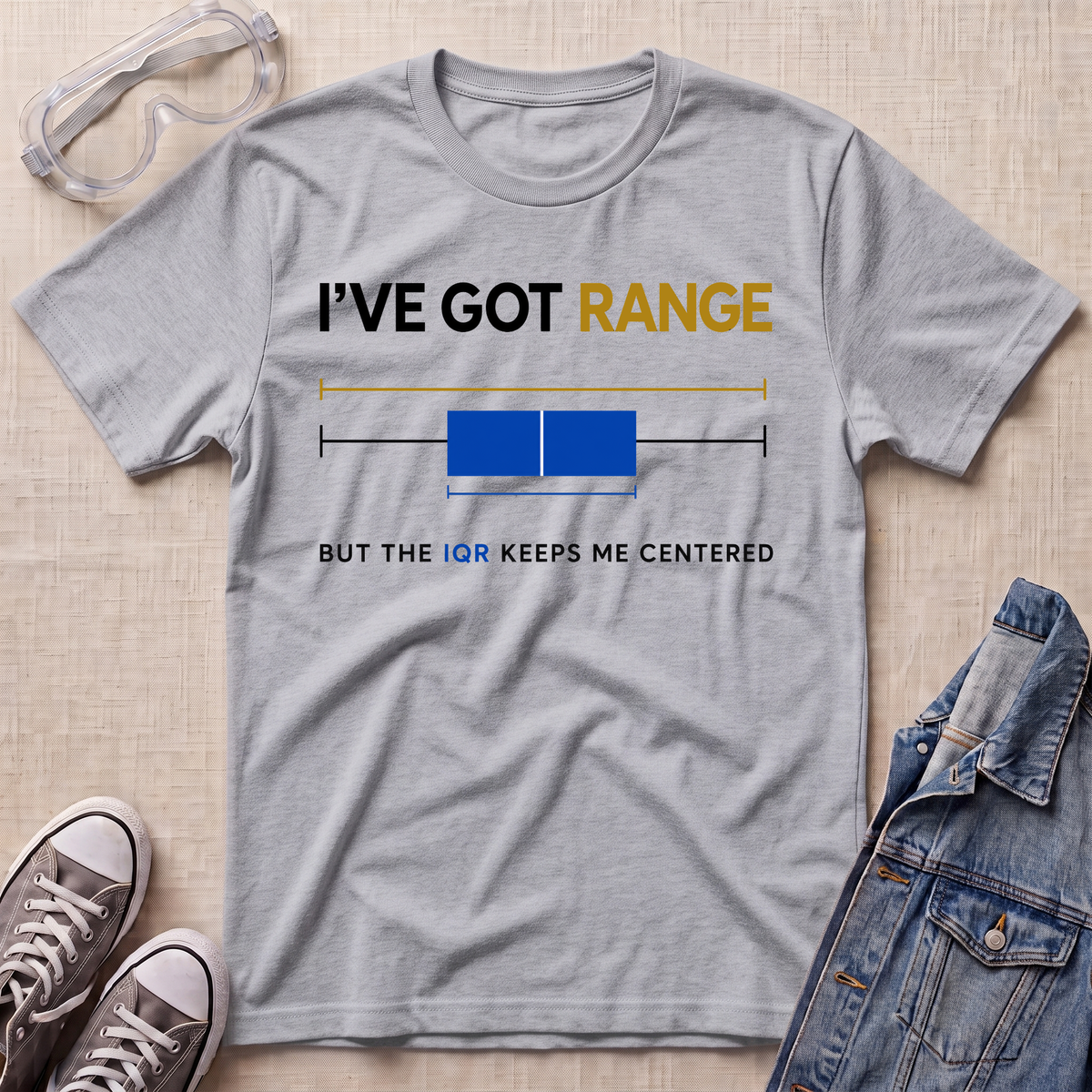 A heather gray graphic t-shirt laid flat on a light wood background, styled with safety goggles, sneakers, and a denim jacket. The t-shirt features a humorous graphic with the text 'I'VE GOT RANGE BUT THE IQR KEEPS ME CENTERED' and a horizontal box plot illustration.