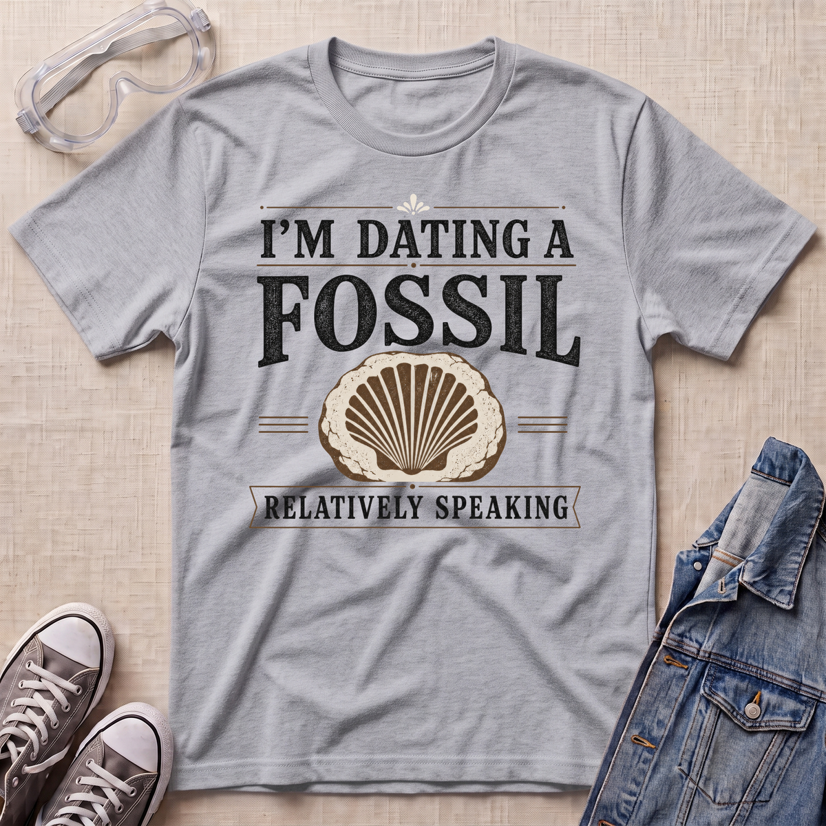 A heather gray short-sleeve t-shirt with a vintage-style graphic that reads 'I'M DATING A FOSSIL RELATIVELY SPEAKING' with a seashell illustration. The shirt is displayed in a flat-lay arrangement on a beige textured background, styled with safety goggles, sneakers, and a denim jacket.