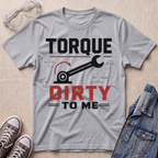 A heather gray graphic t-shirt laid flat on a light wood background, styled with safety glasses, gray sneakers, and a denim jacket. The t-shirt features a black and red graphic that reads 'TORQUE DIRTY TO ME' with a wrench and gear illustration.