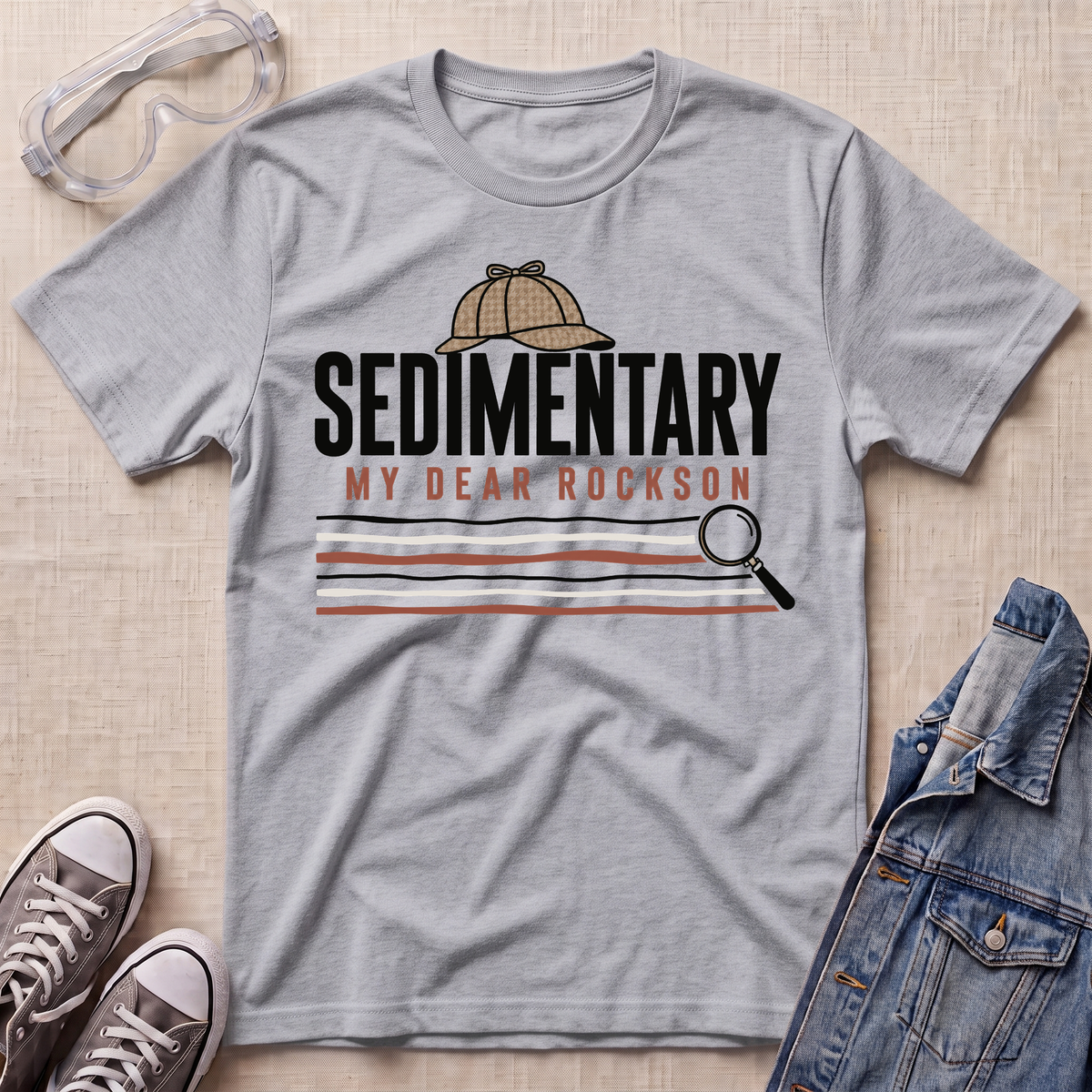 A heather grey graphic t-shirt featuring a detective themed 'Sedimentary My Dear Rockson' print, styled in a flat-lay with a denim jacket, sneakers, and safety goggles on a textured beige background.