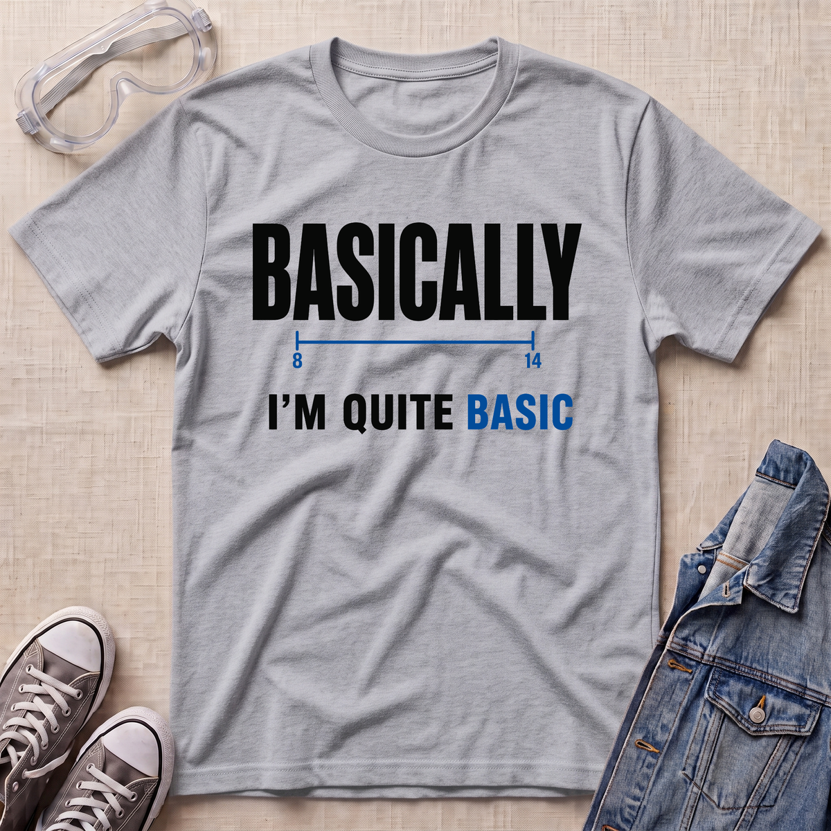 A heather gray graphic t-shirt laid flat on a light beige textured background, styled with safety goggles, gray sneakers, and a denim jacket. The t-shirt features the text 'BASICALLY I'M QUITE BASIC' in black and blue lettering.