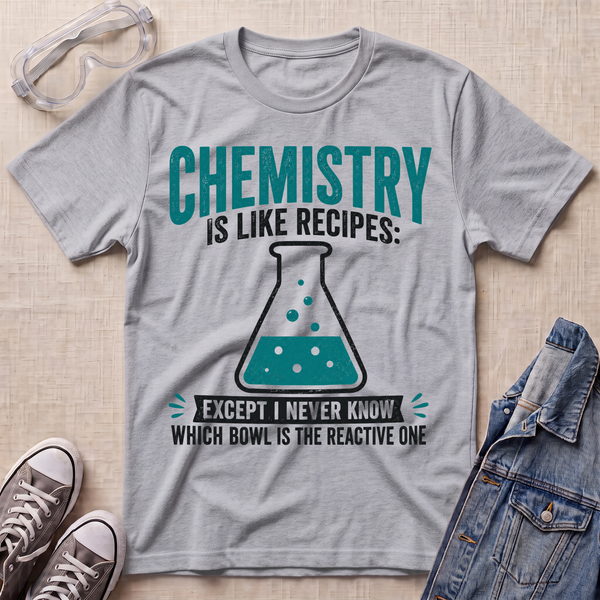 A heather gray graphic t-shirt laid flat on a beige fabric background, styled with safety goggles, gray sneakers, and a denim jacket. The t-shirt features a teal and black graphic that reads 'CHEMISTRY IS LIKE RECIPES: EXCEPT I NEVER KNOW WHICH BOWL IS THE REACTIVE ONE' with an illustration of a beaker.