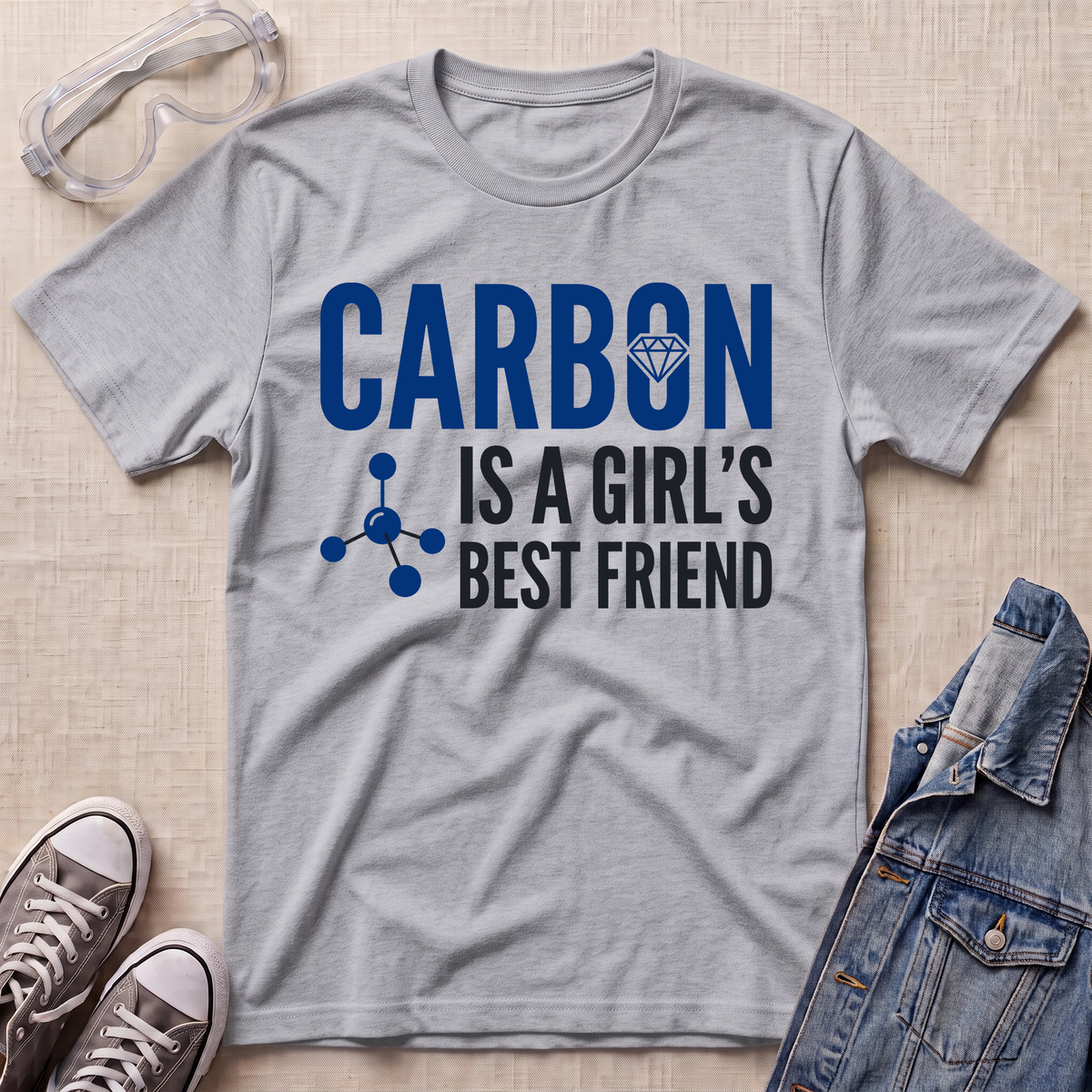 A heather gray graphic t-shirt laid flat on a beige fabric background, surrounded by styling props including safety goggles, gray sneakers, and a denim jacket. The t-shirt features the text 'CARBON IS A GIRL'S BEST FRIEND' in blue and white, with a small blue molecule graphic.
