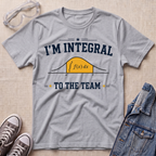 A heather gray graphic t-shirt laid flat on a light wood background, styled with safety glasses, gray sneakers, and a denim jacket. The t-shirt features a navy blue and yellow graphic that reads 'I'M INTEGRAL TO THE TEAM' with a simple integral curve illustration.