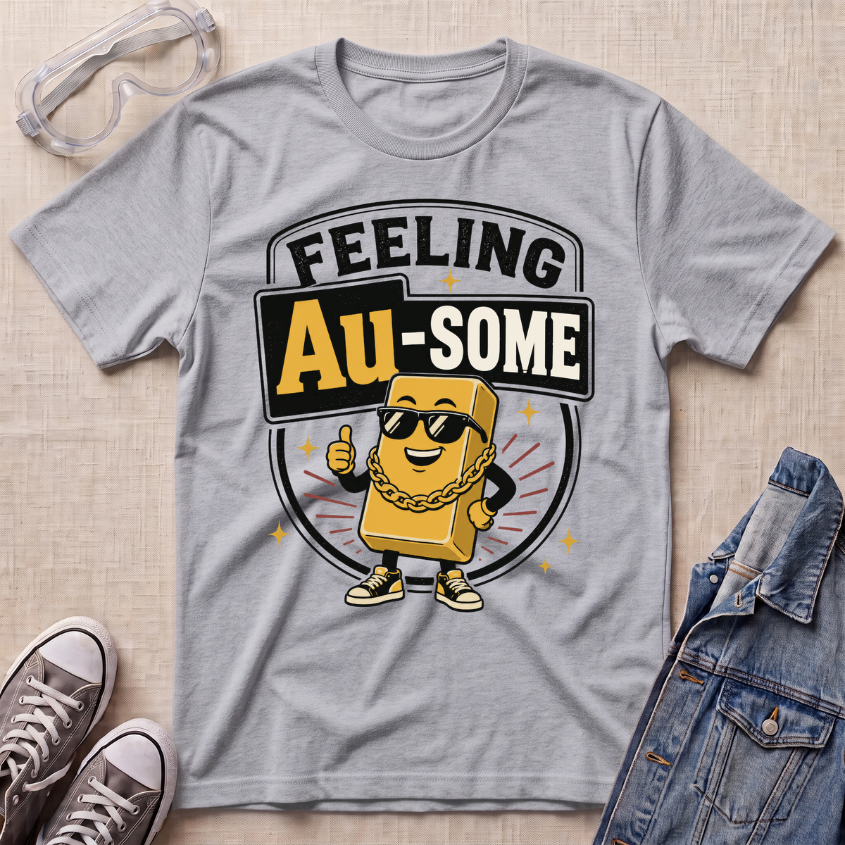 A heather gray graphic t-shirt featuring a cartoon gold bar character wearing sunglasses and a gold chain with the text 'FEELING Au-SOME'. The t-shirt is styled in a flat-lay arrangement on a beige textured background, surrounded by safety goggles, gray sneakers, and a denim jacket.