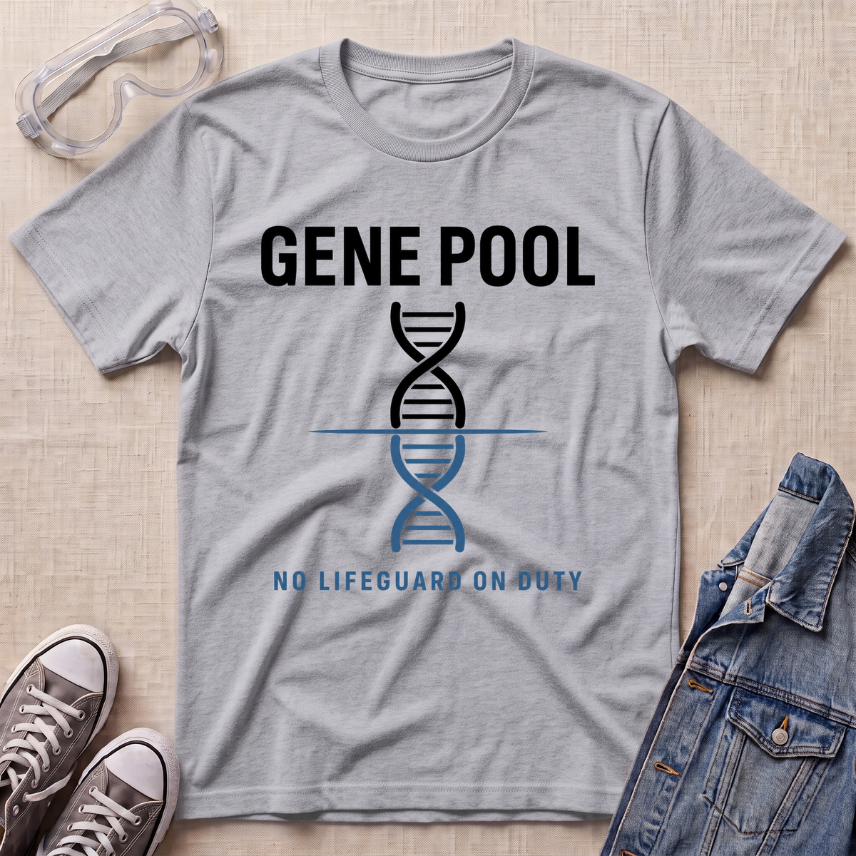 A heather gray short-sleeve t-shirt laid flat on a beige textured surface, featuring a black and blue DNA double helix graphic with the text 'GENE POOL' and 'NO LIFEGUARD ON DUTY'. Goggles, sneakers, and a denim jacket are styled around the shirt as props.