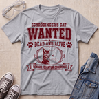 A heather gray graphic T-shirt laid flat on a beige textured surface, styled with safety goggles, sneakers, and a denim jacket. The shirt features a maroon 'WANTED' graphic for Schrödinger's Cat with a cat illustration and paw prints.