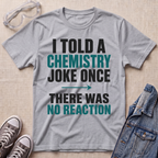A heather gray graphic t-shirt laid flat on a light wood background, surrounded by styling props including safety goggles, sneakers, and a denim jacket. The shirt features a bold text graphic that reads 'I TOLD A CHEMISTRY JOKE ONCE THERE WAS NO REACTION' in black and teal lettering.