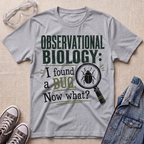 A heather gray graphic t-shirt laid flat on a beige textured surface, surrounded by styling props including clear safety goggles, gray sneakers, and a denim jacket. The t-shirt features a dark green and black graphic that reads 'OBSERVATIONAL BIOLOGY: I found a BUG.