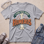 A heather gray graphic t-shirt laid flat on a light wood surface, featuring a bone graphic and the text 'I FIND THIS SHIRT HUMERUS' in orange and green. The shirt is styled with safety glasses, gray sneakers, and a denim jacket.