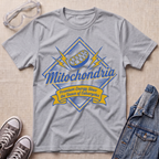 A heather gray short-sleeve t-shirt with a blue and yellow 'Mitochondria' graphic, styled in a flat lay with safety goggles, sneakers, and a denim jacket on a beige textured background.