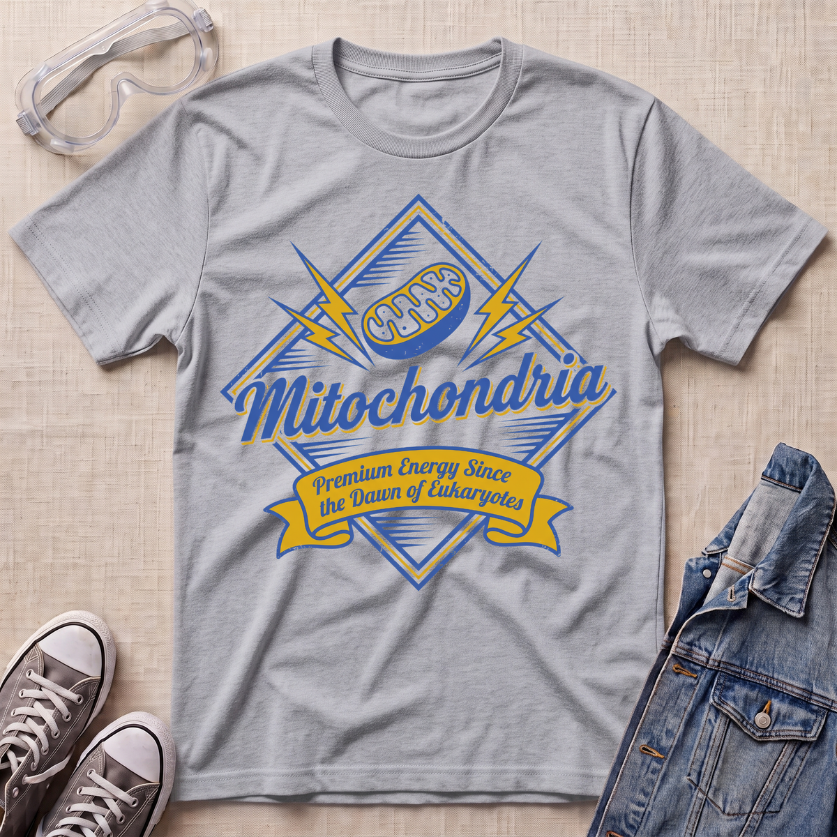 A heather gray short-sleeve t-shirt with a blue and yellow 'Mitochondria' graphic, styled in a flat lay with safety goggles, sneakers, and a denim jacket on a beige textured background.