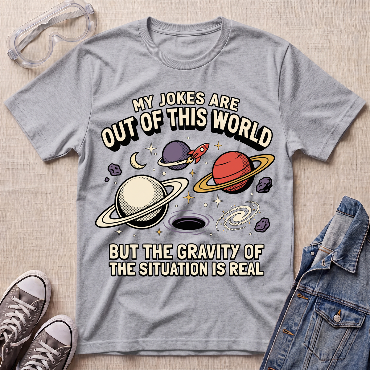 A heather sport gray graphic t-shirt featuring a space-themed design with planets, stars, and the text 'MY JOKES ARE OUT OF THIS WORLD BUT THE GRAVITY OF THE SITUATION IS REAL'. The shirt is displayed in a flat-lay arrangement on a beige textured background, surrounded by props including safety glasses, gray sneakers, and a denim jacket.