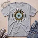 A heather gray short-sleeve t-shirt with a circular graphic reading 'OFFICIAL MEMBER THE HEISENBERG UNCERTAINTY PRINCIPLE FAN CLUB' and an atom symbol, styled in a flat lay with safety goggles, sneakers, and a denim jacket on a beige textured background.