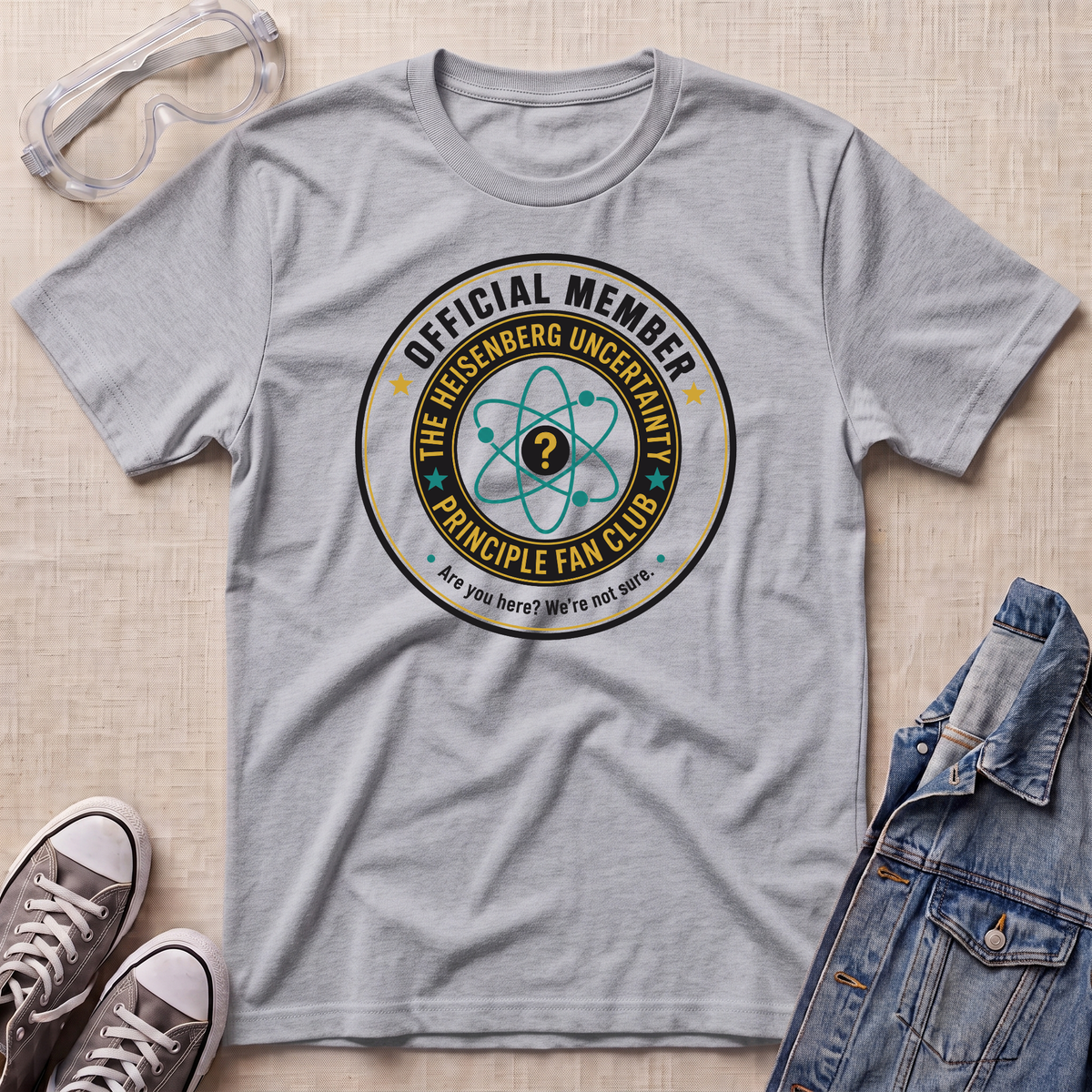 A heather gray short-sleeve t-shirt with a circular graphic reading 'OFFICIAL MEMBER THE HEISENBERG UNCERTAINTY PRINCIPLE FAN CLUB' and an atom symbol, styled in a flat lay with safety goggles, sneakers, and a denim jacket on a beige textured background.