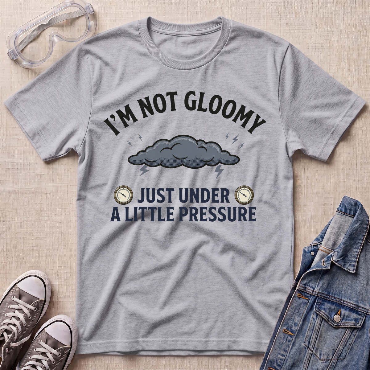 A heather gray graphic t-shirt laid flat on a light beige textured surface, styled with safety goggles, sneakers, and a denim jacket. The t-shirt features a dark gray cloud graphic with rain and the text 'I'M NOT GLOOMY JUST UNDER A LITTLE PRESSURE'.