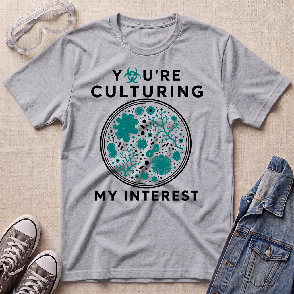 A heather gray graphic t-shirt laid flat on a light beige textured surface, surrounded by styling props including safety goggles, sneakers, and a denim jacket. The t-shirt features a circular petri-dish style graphic with teal microorganisms and the text 'YOU'RE CULTURING MY INTEREST' in black.