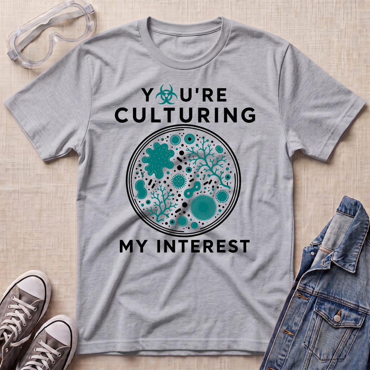 A heather gray graphic t-shirt laid flat on a light beige textured surface, surrounded by styling props including safety goggles, sneakers, and a denim jacket. The t-shirt features a circular petri-dish style graphic with teal microorganisms and the text 'YOU'RE CULTURING MY INTEREST' in black.