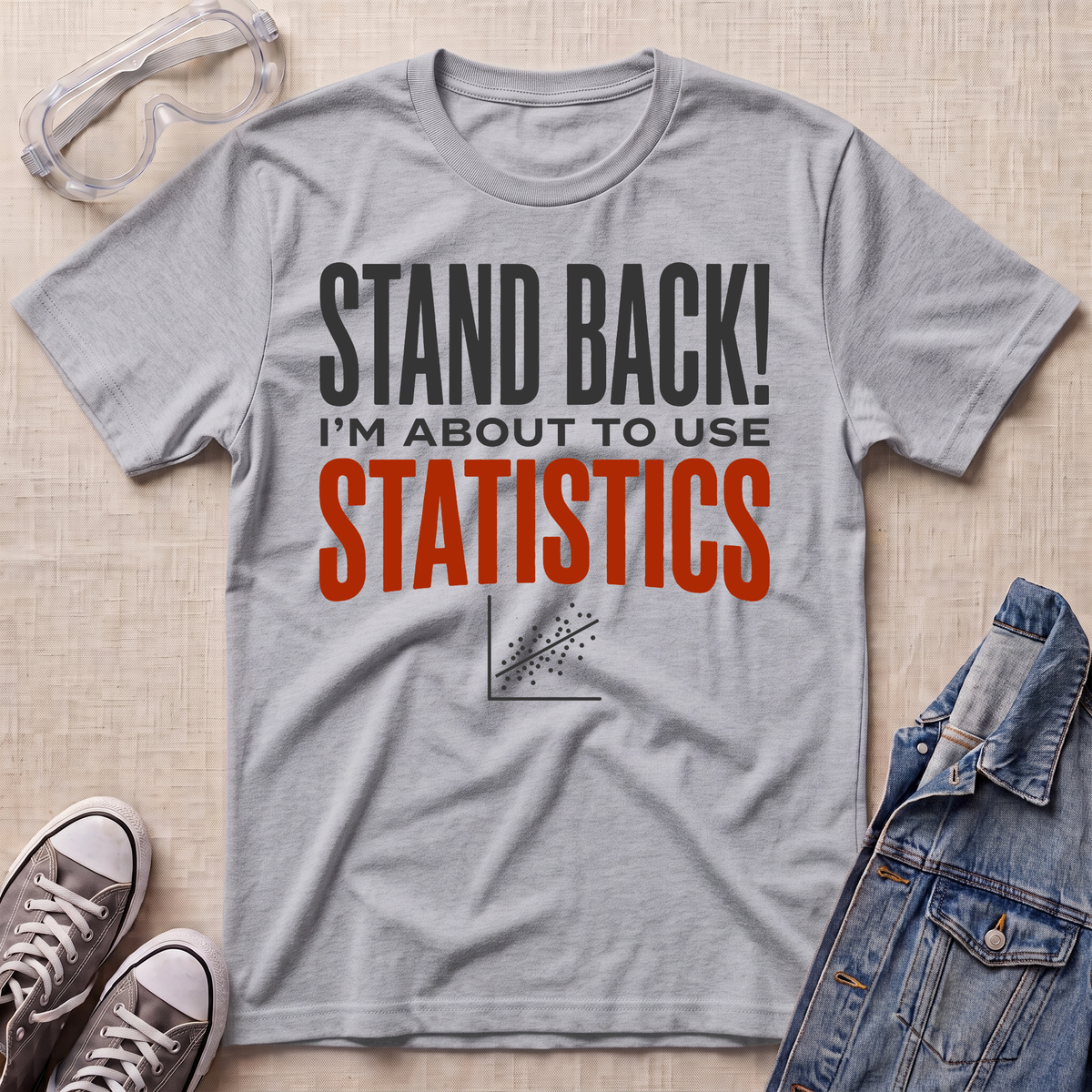 A heather gray graphic t-shirt laid flat on a light wood background, styled with safety goggles, sneakers, and a denim jacket. The t-shirt features the text 'STAND BACK! I'M ABOUT TO USE STATISTICS' in black and orange lettering.