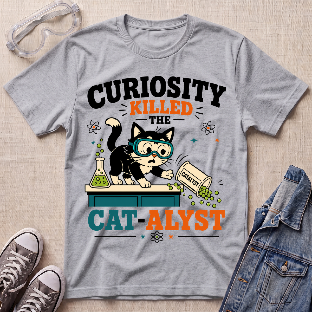 A heather gray graphic T-shirt laid flat on a light fabric surface, surrounded by styling props including safety goggles, sneakers, and a denim jacket. The shirt features a cartoon black-and-white cat wearing goggles and pouring green powder from a labeled bottle, with the text “CURIOSITY KILLED THE CAT-ALYST.