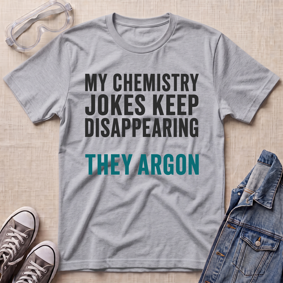A heather gray graphic t-shirt with the text 'MY CHEMISTRY JOKES KEEP DISAPPEARING THEY ARGON' in white and teal lettering, styled in a flat lay with safety goggles, gray sneakers, and a denim jacket on a beige fabric background.