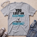 A heather gray graphic t-shirt laid flat on a beige textured surface, surrounded by styling props including clear safety goggles, gray sneakers, and a denim jacket. The t-shirt features a black and teal graphic that reads 'YOU LOST AN ELECTRON? ARE YOU POSITIVE?' with a simple electron illustration.
