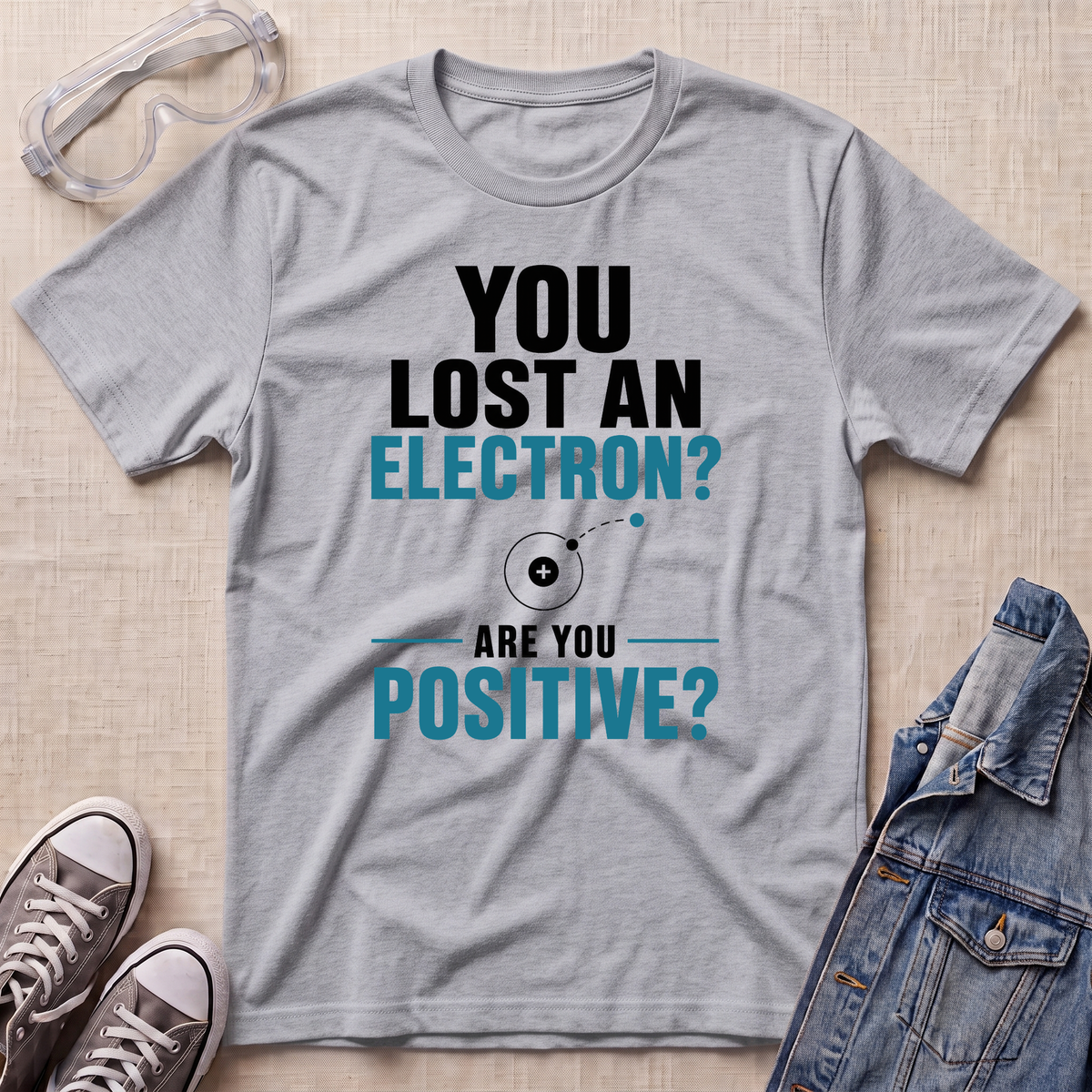A heather gray graphic t-shirt laid flat on a beige textured surface, surrounded by styling props including clear safety goggles, gray sneakers, and a denim jacket. The t-shirt features a black and teal graphic that reads 'YOU LOST AN ELECTRON? ARE YOU POSITIVE?' with a simple electron illustration.