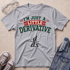 A heather gray graphic t-shirt laid flat on a light wood background, styled with safety glasses, gray sneakers, and a denim jacket. The t-shirt features the text 'I'M JUST A LITTLE DERIVATIVE' in green and red, with a black 'd/dx' formula printed below.