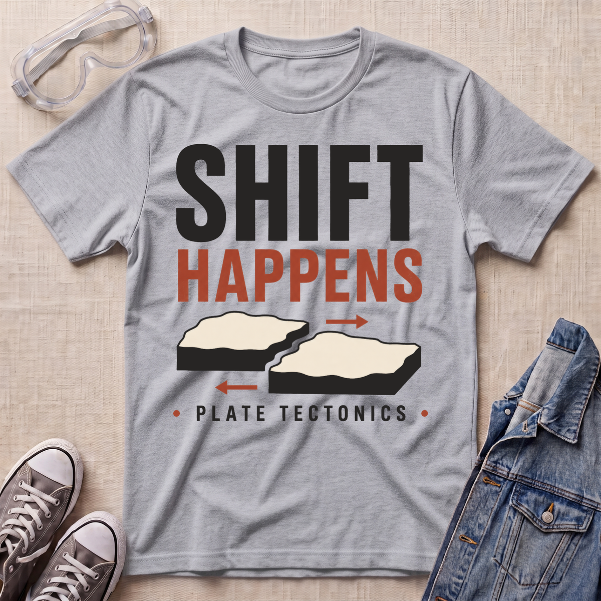 A heather gray graphic t-shirt laid flat on a beige textured surface, styled with safety goggles, sneakers, and a denim jacket. The t-shirt features a graphic that reads 'SHIFT HAPPENS' in black and orange, with illustrations of two splitting tectonic plates and the text 'PLATE TECTONICS' below.