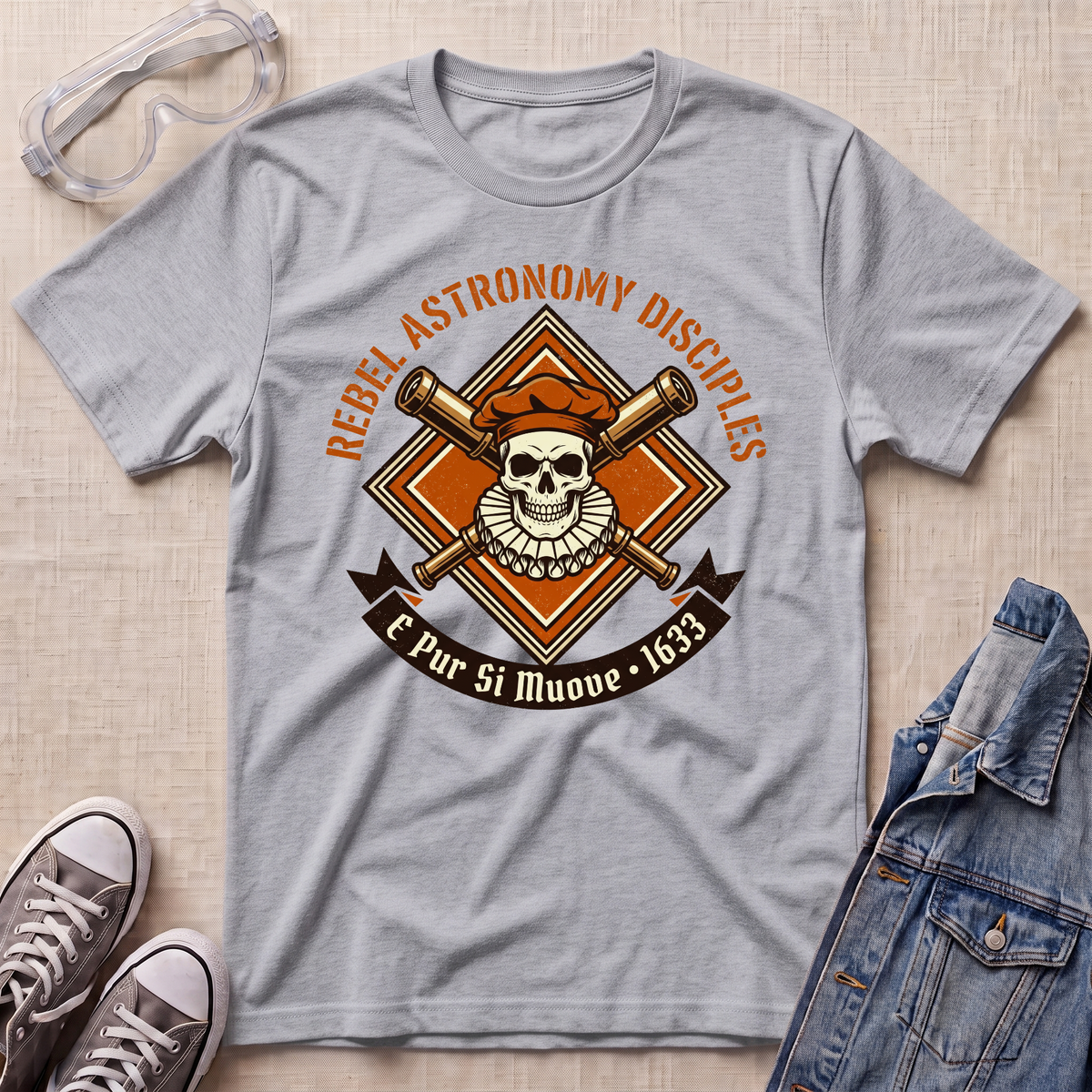 A heather gray graphic t-shirt laid flat on a textured beige surface, styled with safety goggles, gray sneakers, and a denim jacket. The t-shirt features a retro-style astronomy-themed graphic with a skull, crossed telescopes, and the text 'REBEL ASTRONOMY DISCIPLES' and 'E Pur Si Muove - 1633'.