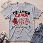 A heather gray graphic t-shirt laid flat on a beige textured surface, surrounded by props including clear safety goggles, gray sneakers, and a blue denim jacket. The t-shirt features a cartoon graphic of a red heart, a pink brain, and a pink stomach with the text 'ORGANIZED CHAOS' and 'HUMAN BODY DEPT.