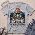 A heather gray graphic t-shirt laid flat on a beige textured background, surrounded by lifestyle props including safety goggles, sneakers, and a denim jacket. The t-shirt features a retro-style graphic of a gneiss rock wearing sunglasses in front of mountains and pine trees, with the text 'STILL GNEISS UNDER PRESSURE'.
