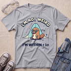 A heather gray graphic t-shirt featuring a platypus wearing glasses under a blue bell curve with the text 'I'M NOT WEIRD' and 'I'M WITHIN ±1σ'. The t-shirt is styled in a flat-lay arrangement on a textured beige background, surrounded by props including safety goggles, gray sneakers, and a denim jacket.