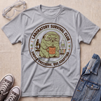 A heather gray graphic t-shirt laid flat on a beige textured surface, surrounded by styling props including safety goggles, gray sneakers, and a denim jacket. The t-shirt features a circular graphic of a green cartoon creature wearing goggles and a mask, holding a mug, with the text 'LABORATORY SURVIVAL CLUB' and 'UNDER PRESSURE, BUT ADAPTING'.