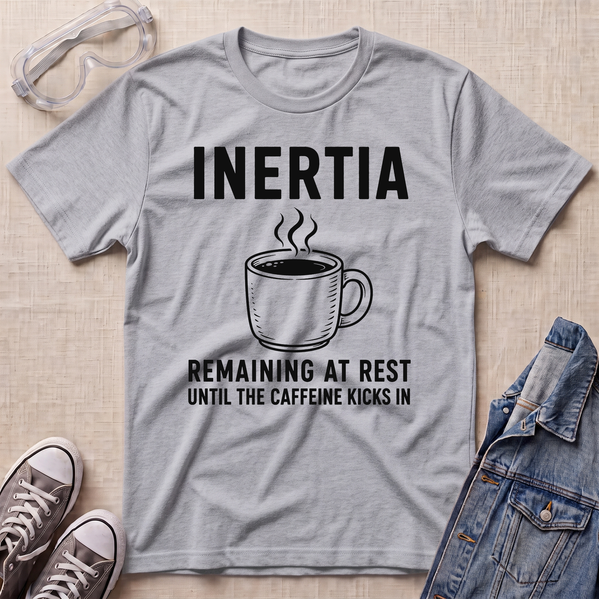 A heather gray graphic t-shirt laid flat on a beige textured background, surrounded by styling props including safety goggles, sneakers, and a denim jacket. The t-shirt features a black graphic of a steaming coffee mug with the text 'INERTIA REMAINING AT REST UNTIL THE CAFFEINE KICKS IN'.