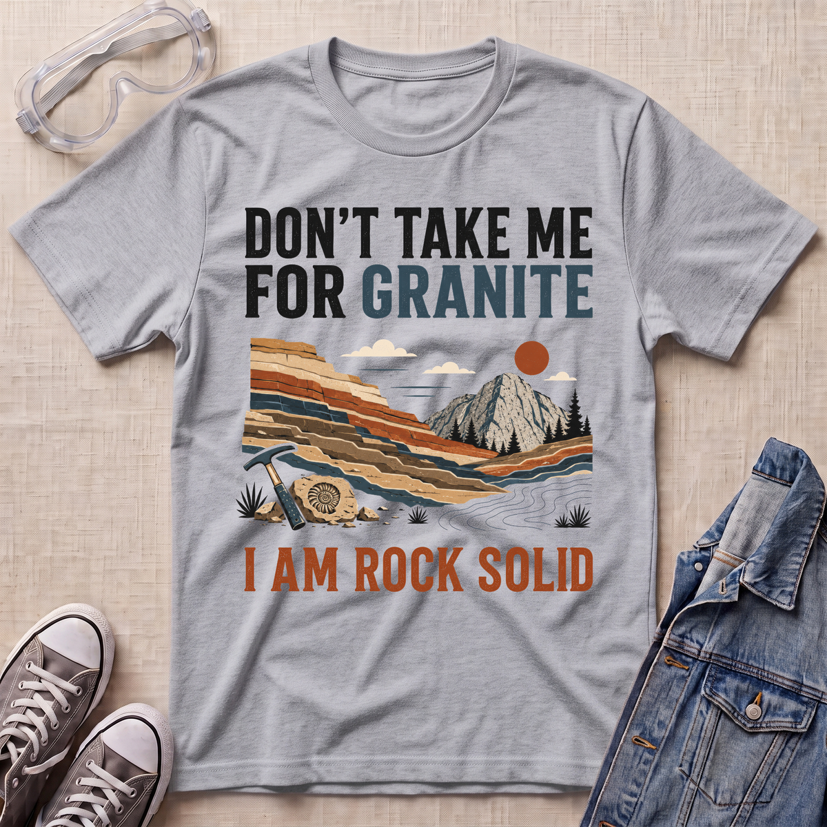 A heather gray graphic t-shirt laid flat on a beige textured surface, surrounded by styling props including safety goggles, sneakers, and a denim jacket. The t-shirt features a retro landscape graphic with the text 'DON'T TAKE ME FOR GRANITE' and 'I AM ROCK SOLID'.