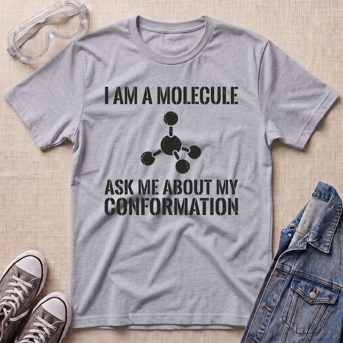 A heather gray short-sleeve t-shirt with a black printed graphic and text reading 'I AM A MOLECULE ASK ME ABOUT MY CONFORMATION' with a simple molecule model. The shirt is displayed in a flat-lay arrangement on a beige textured background, styled with safety goggles, sneakers, and a denim jacket.