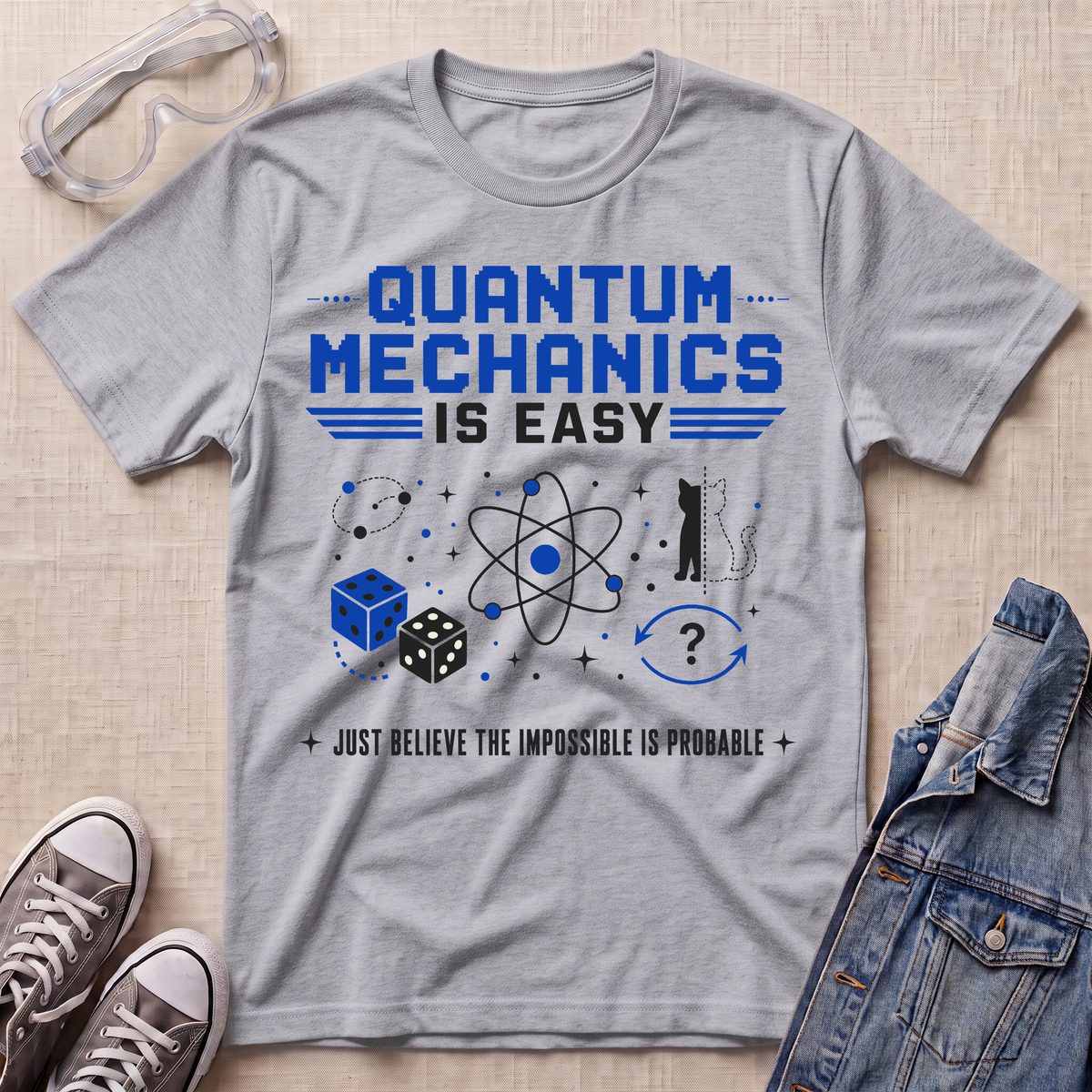 A heather gray short-sleeve t-shirt with a blue and black graphic reading 'QUANTUM MECHANICS IS EASY' and 'JUST BELIEVE THE IMPOSSIBLE IS PROBLEMA', styled in a flat lay with safety goggles, sneakers, and a denim jacket on a beige textured background.