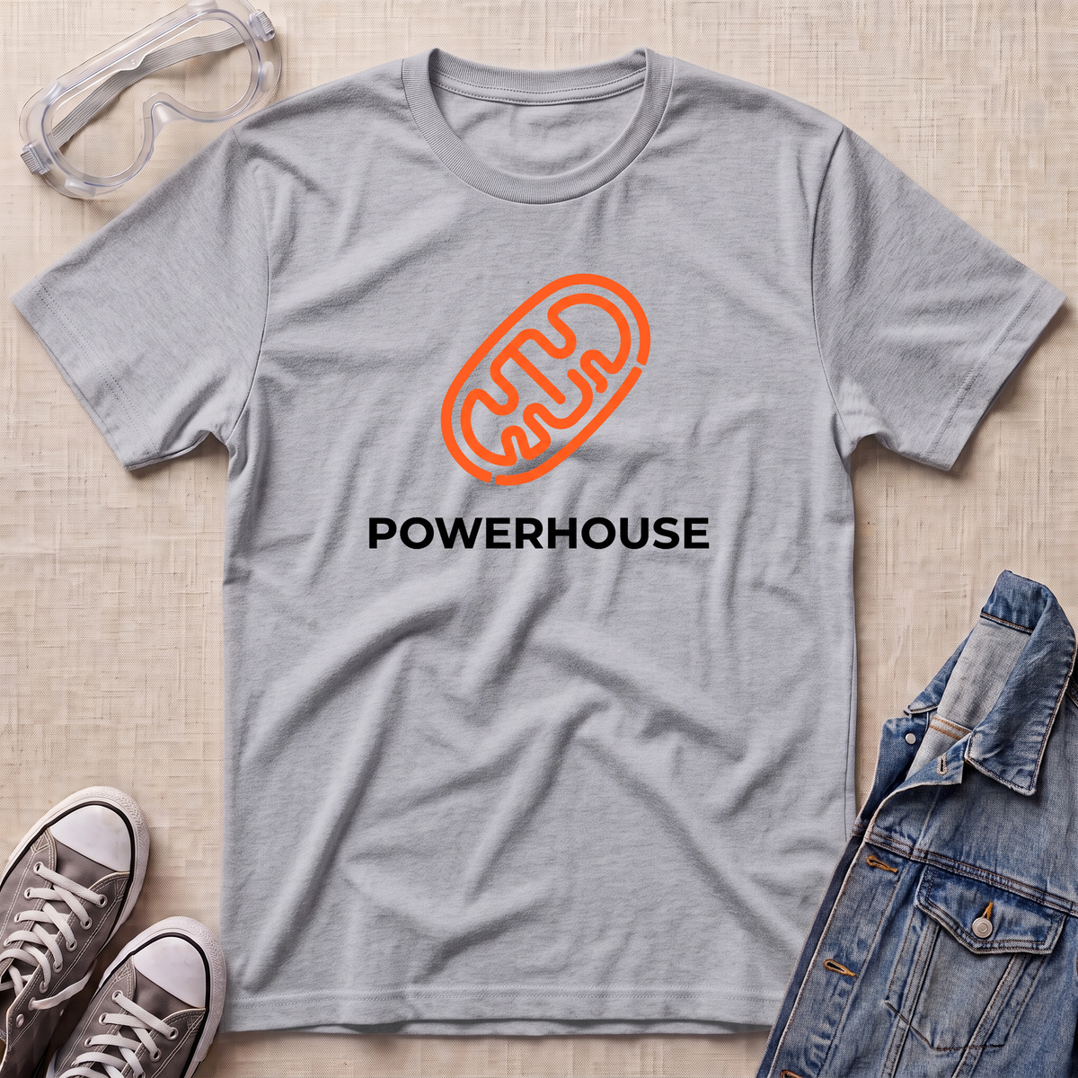 A heather gray short-sleeve t-shirt with an orange mitochondrial outline and the word 'POWERHOUSE' printed in black on the chest, styled in a flat lay with safety goggles, sneakers, and a denim jacket on a light beige textured background.