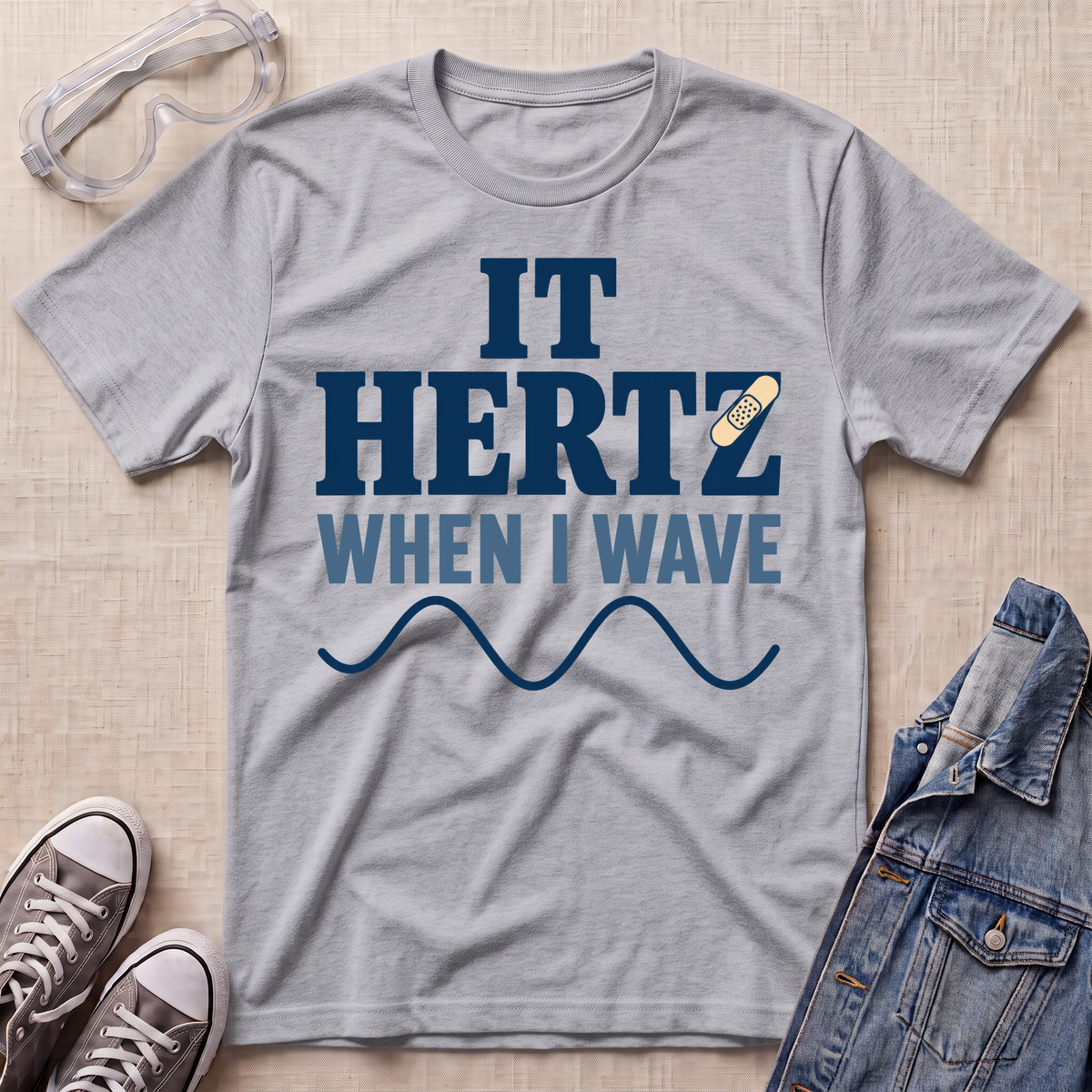 A heather gray graphic t-shirt laid flat on a light wood background, styled with safety goggles, sneakers, and a denim jacket. The t-shirt features the text 'IT HERTZ WHEN I WAVE' in navy blue and light blue, with a small band-aid graphic and a wavy line below.