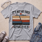 A heather gray graphic t-shirt laid flat on a textured beige fabric background, styled with safety glasses, gray sneakers, and a blue denim jacket. The t-shirt features a humorous graphic with the text 'IT'S NOT MY FAULT ACTUALLY, IT IS' surrounding a retro cross-section illustration of a desert landscape.