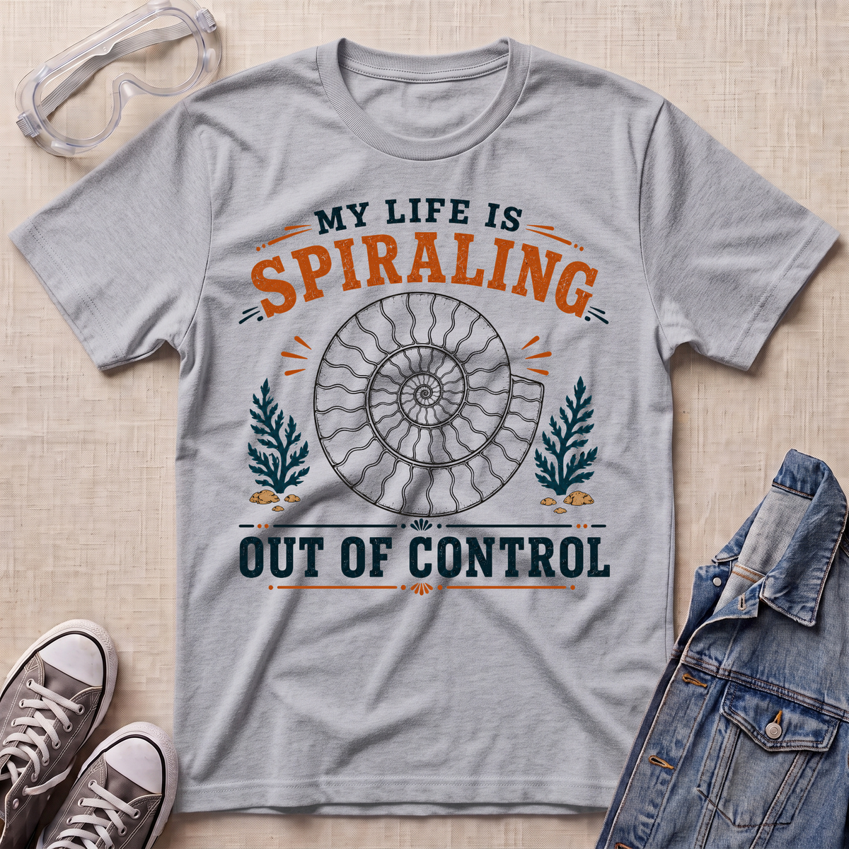 A heather gray graphic t-shirt laid flat on a beige textured surface, surrounded by styling props including safety glasses, gray sneakers, and a denim jacket. The t-shirt features a retro-style graphic with a spiral ammonite shell and the text 'MY LIFE IS SPIRALING OUT OF CONTROL' in orange and dark teal.